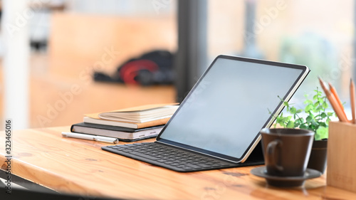 Photo of Computer tablet with white blank screen in keyboard case putting together with stack of notebook, potted plant, coffee cup, pencil holder on the modern wooden table with blurred background.