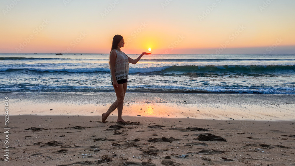 A girl holding a setting sun in her hand on Seminyak beach on Bali, Indonesia. The sun sets directly into the water. Calm sea washes gently the shore. The sunbeams reflecting on the sea surface. Fun