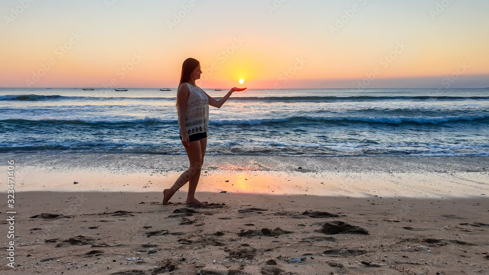 A girl holding a setting sun in her hand on Seminyak beach on Bali, Indonesia. The sun sets directly into the water. Calm sea washes gently the shore. The sunbeams reflecting on the sea surface. Fun A girl holding a setting sun in her hand on Seminyak beach on Bali, Indonesia. The sun sets directly into the water. Calm sea washes gently the shore. The sunbeams reflecting on the sea surface. Fun