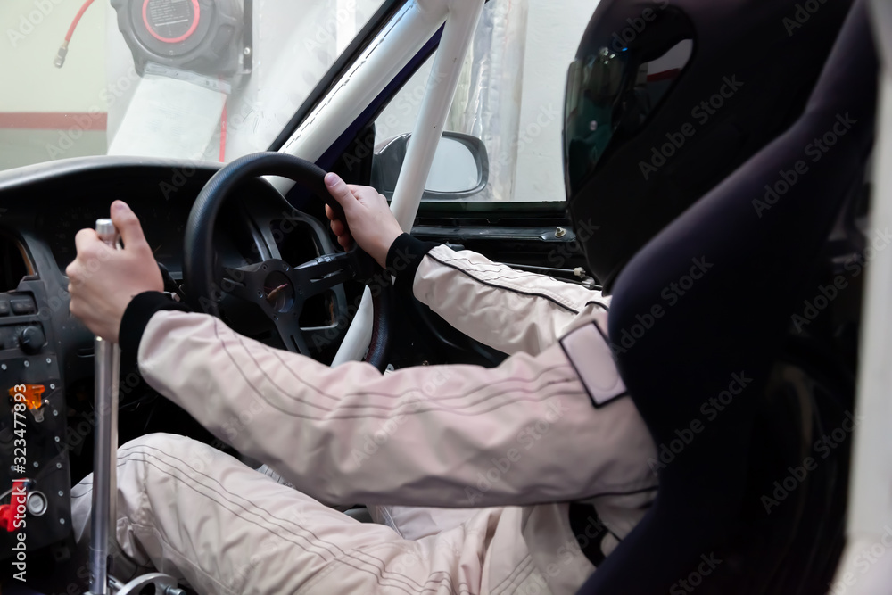 A professional racer in a black helmet and a white homologated suit sits in the sports seat of a car with a roll cage for drifting and racing during race and training.