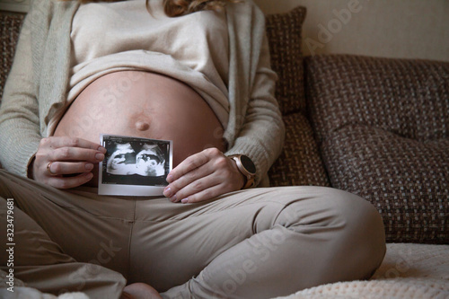 Cropped shot of pregnant woman holding ultrasaund picture on her  tummy at home interiors. Close-up female hands keeping scan. Maternity prenatal concept