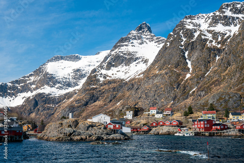 The city of Aa in Lofoten, Norway. Tiny red houses called rorbuer underneath steep snow covered mountain peaks and blue sky. Traveling and explorer concept.