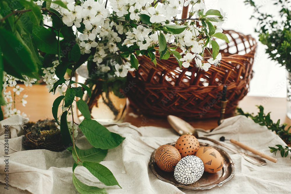 Fototapeta premium Natural Easter eggs painted with wax on vintage plate on background of wicker basket, white spring flowers and greenery on wooden table. Happy Easter. Stylish Rural still life. Zero waste