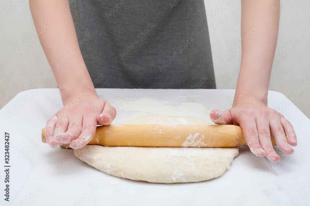 Hands roll out the dough with a rolling pin, on a white background, on the table. The concept is to make the dough, pizza, baking recipes. Hands roll out the dough with a rolling pin, on a white background, on the table. The concept is to make the dough, pizza, baking recipes.