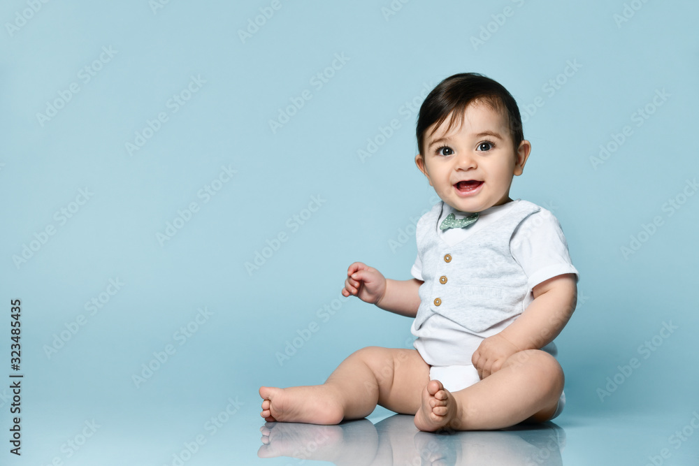 Little child in white bodysuit as vest with bow-tie, barefoot. He smiling, sitting on the floor against blue background. Close up Little child in white bodysuit as vest with bow-tie, barefoot. He smiling, sitting on the floor against blue background. Close up