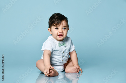 Baby boy in white bodysuit as a vest with bow-tie, barefoot. He smiling, sitting on the floor against blue background. Close up