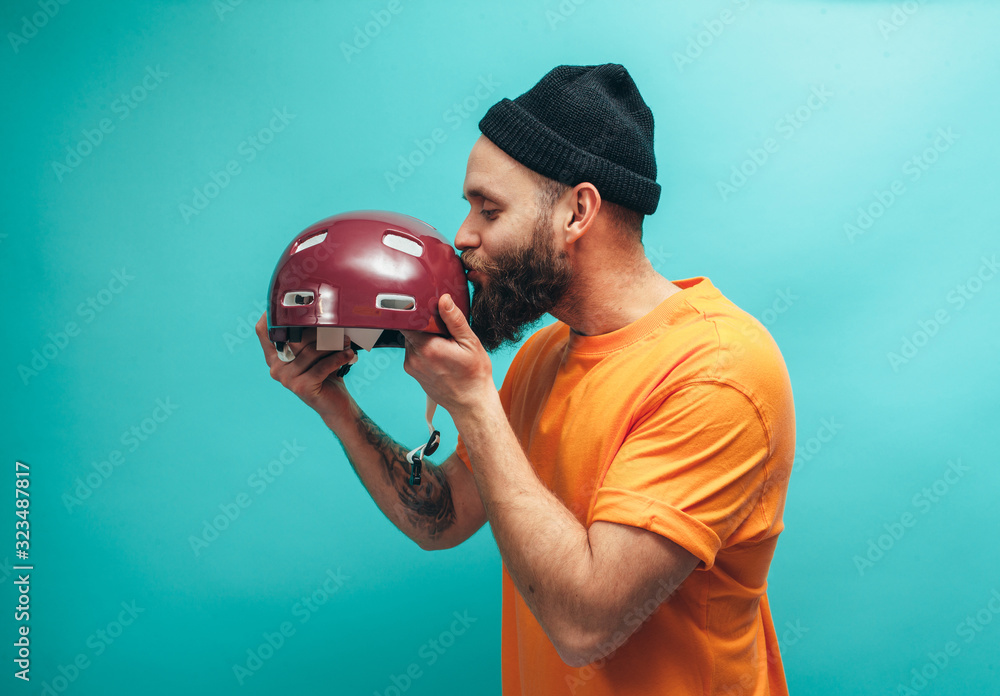 Portrait of handsome hipster guy with beard wearing blank orange t-shirt and red bike helmet , isolated on blue studio wall.