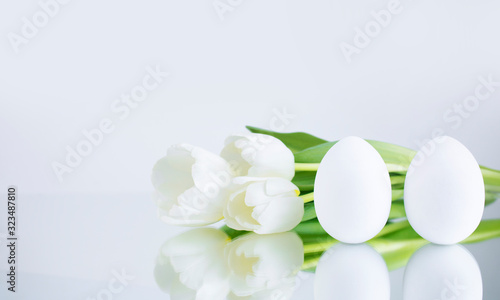 Chicken eggs  painted white on a gray background  are reflected on the mirror surface  next to the bouquet  with white tulips. Creative card with Easter. Banner. Copy space