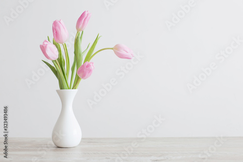 pink tulips in white ceramic vase on wooden table on background white wall #323493486