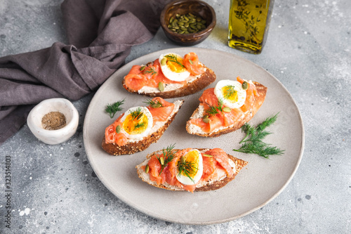 Italian appetizer bruschetta with red fish, boiled egg, cream cheese and dill on a grey plate on a grey background, close up horizontal