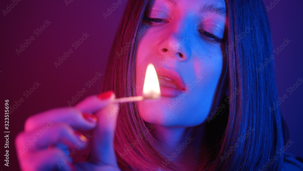 Obraz premium Attractive young woman holding matchstick with light in fingers, blowing out, looking at camera. Blue and purple combination of lights. Bob hairstyle. Red nails.