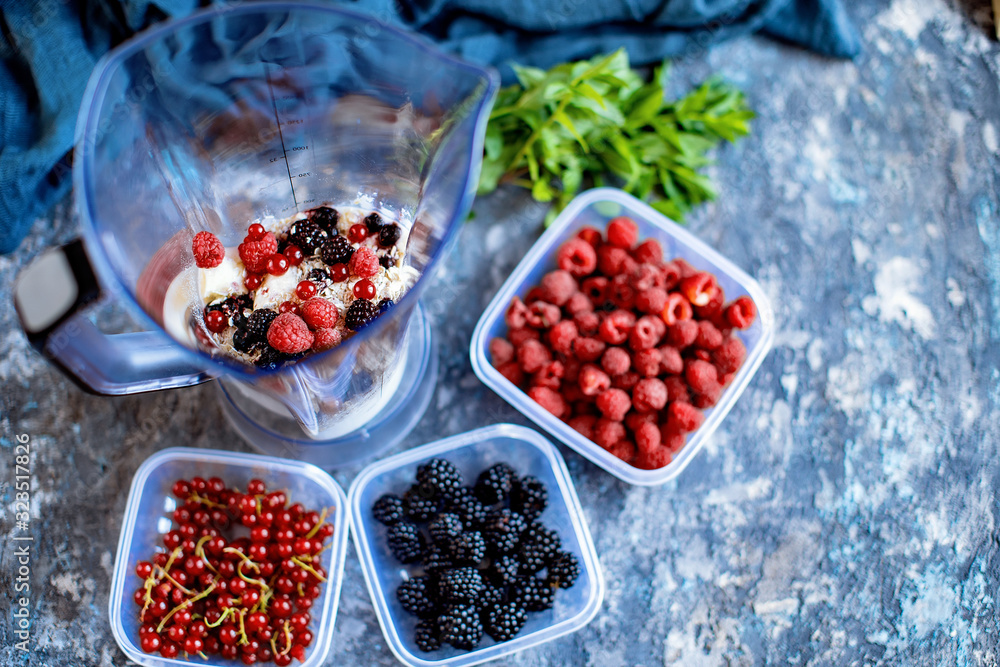 Blender with oatmeal, plastic plates, containers with raspberry, blackberry, red currant, cherry on a stone background. Making a healthy smoothie on a blender. Blender with oatmeal, plastic plates, containers with raspberry, blackberry, red currant, cherry on a stone background. Making a healthy smoothie on a blender.