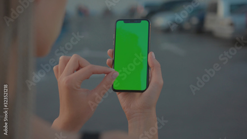 Lviv, Ukraine - May 19, 2018: Close-up of young urban girl showing vertical smartphone with greenscreen chrome key technology on parking lot outside. City life and modern gadgets.