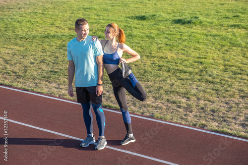 Young couple of fit sportsmen boy and girl doing warming up exercises before running on red tracks of public stadium outdoors.