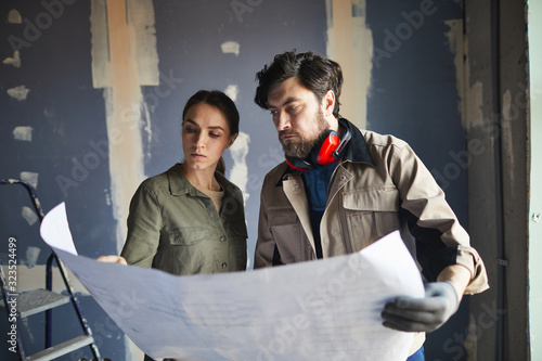 Portrait of young woman looking at floor plans with building contractor while standing against dry wall in under construction house, copy space photo
