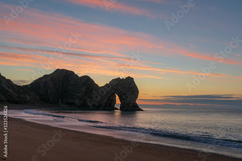 A photograph of Durdle door and crashing waves taken during dawn, with a sunrise of red and amber clouds