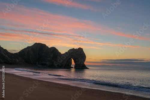 Obraz na płótnie A photograph of Durdle door and crashing waves taken during dawn, with a sunrise of red and amber clouds w salonie