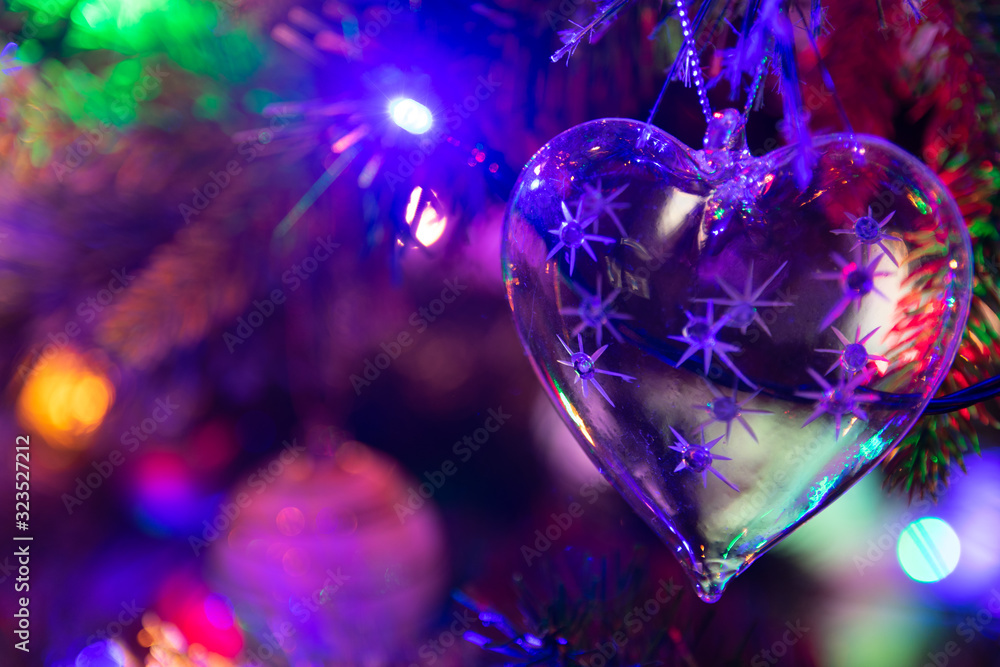 A glass heart shaped Christmas bauble handing from a green Christmas tree surrounded by fairy lights