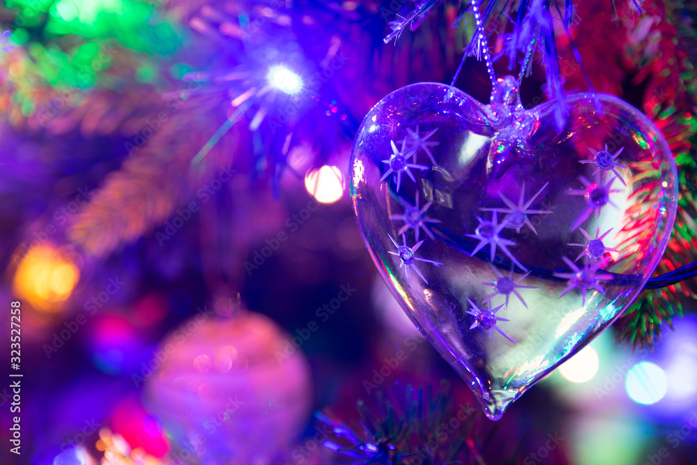 A glass heart shaped Christmas bauble handing from a green Christmas tree surrounded by fairy lights