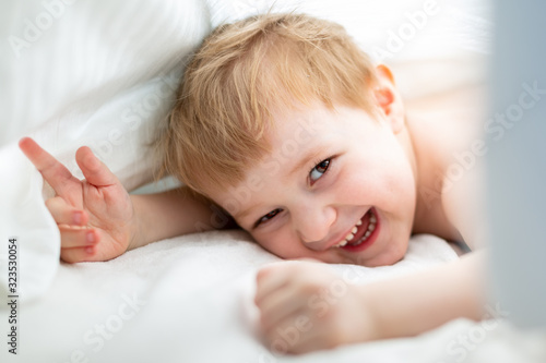 Morning awakening. Cute blonde boy dabbles in white bed. Family having fun at home. Happy kid playing in white bedroom photo