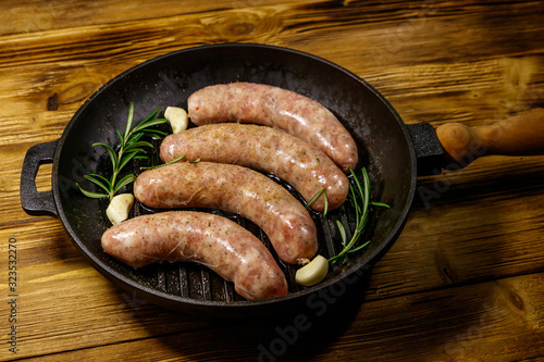 Raw sausages ready for preparation with rosemary, garlic and spices in cast iron grill frying pan on wooden table