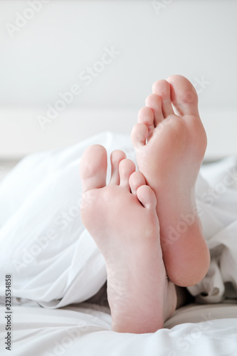 Men's feet under a white blanket, in bed, in a comfortable apartment, in natural light, with copy space. Vertical photo.
