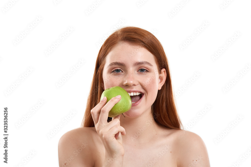 Enjoying healthy lifestyle. Portrait of young and happy red-haired girl biting a fresh green apple and looking at camera while standing against white background Enjoying healthy lifestyle. Portrait of young and happy red-haired girl biting a fresh green apple and looking at camera while standing against white background