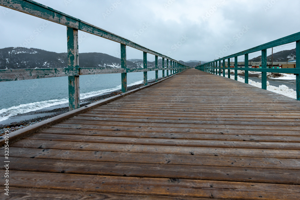 A long wooden jetty near the ocean. The deck is worn textured wood with wooden green rails on both sides. The ocean is on the left and a road on the right side of the boardwalk with buildings.