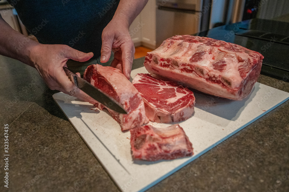 A butcher prepares to cut raw beef prime rib steaks from a large cut of meat. The butcher's hands are using a long stainless steel knife to cut thick steaks and ribs.
