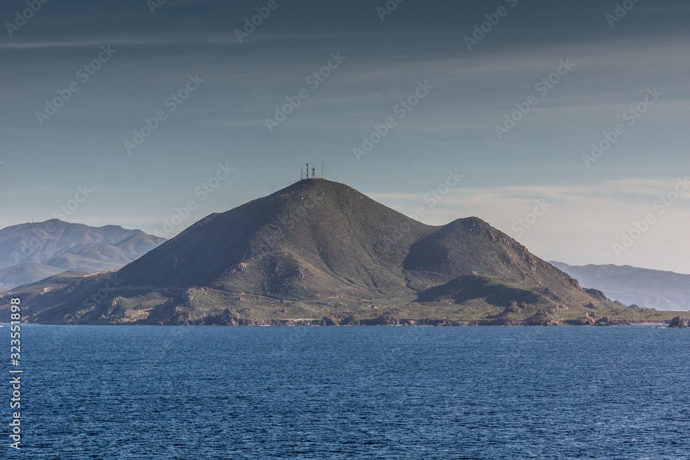 Ensenada, Mexico - January 17, 2012: Dark green mountain with antennas on top behind blue Pacific Ocean under light blue sky at entrance to All Saints Bay.
