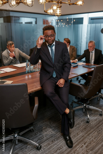 Business professional. Portrait of african businessman in formal wear adjusting his eyeglasses and looking at camera while leaning at the desk in office
