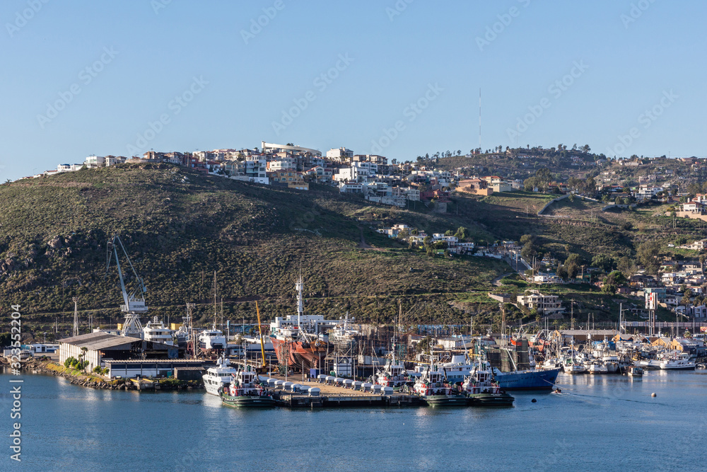 Ensenada, Mexico - January 17, 2012: Several fishing vessels, 1 lifted out of water, are docked at their pier in the port. Sitting on blue bay water and backed by citiscape on hill slope under blue sk