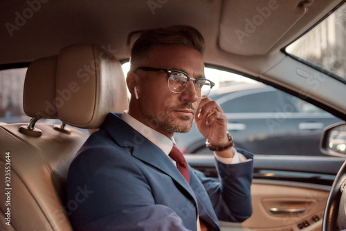Working from everywhere. Handsome mature man in full suit adjusting headphones while driving a car © Svitlana