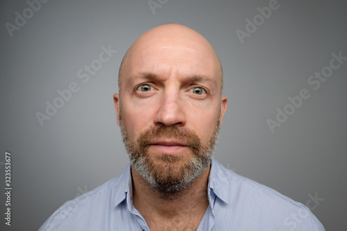 Keep calm and pretend nothing happened. Mature male with beard standing with no emotions over gray background, staring at camera without interest.