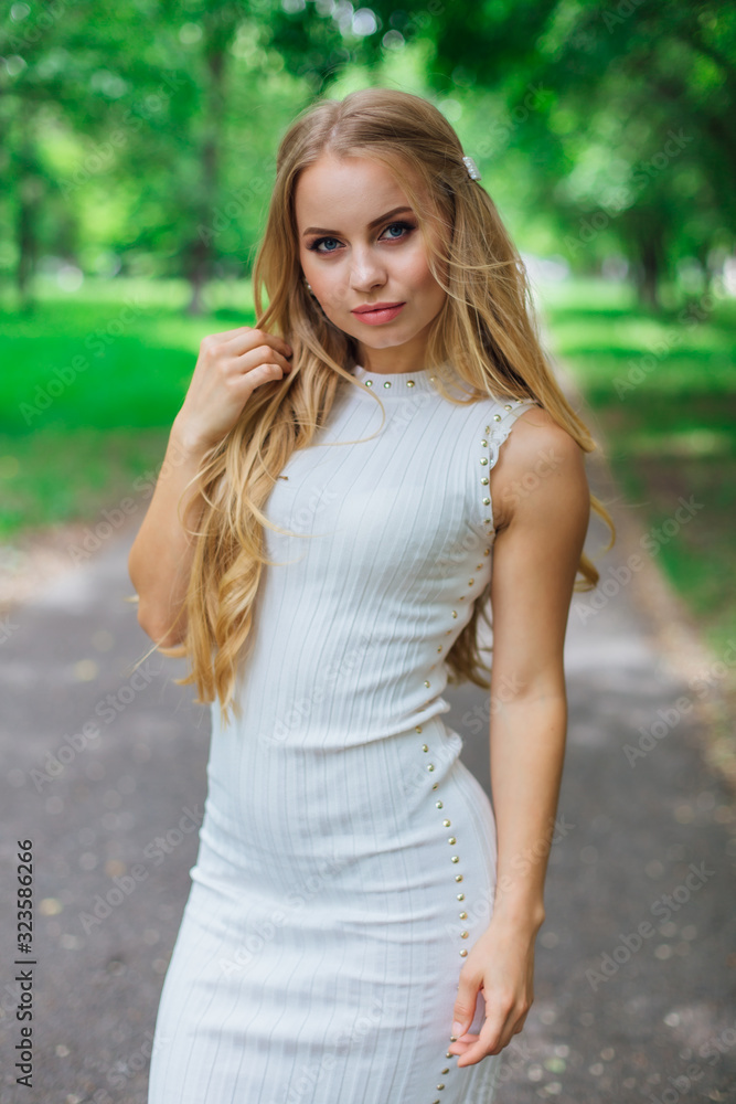Naklejka premium Portrait of a charming blond woman wearing beautiful white dress standing on the road under trees.