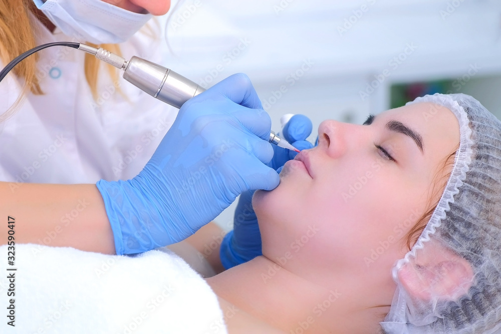 Naklejka premium Cosmetologist is applying permanent makeup on woman lips using tattoo machine in clinic. Beautician and patient face closeup. Lips microblading procedure for girl. Beauty industry concept.