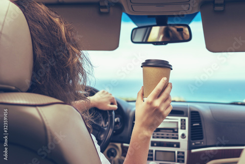 Woman driver drinking coffee paper cup inside car during driving. Girl relaxing in auto trip traveling along ocean tropical beach in background. Traveler concept. Back view