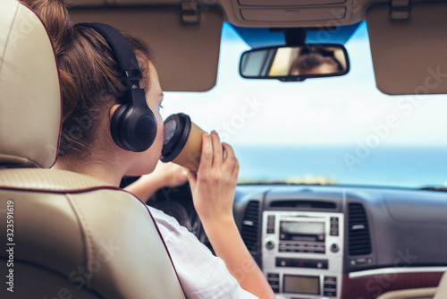 Woman driver in the headphones driving a car. Girl relaxing in auto trip drinking coffee paper cup traveling along ocean tropical beach in background. Traveler concept. © flowertiare
