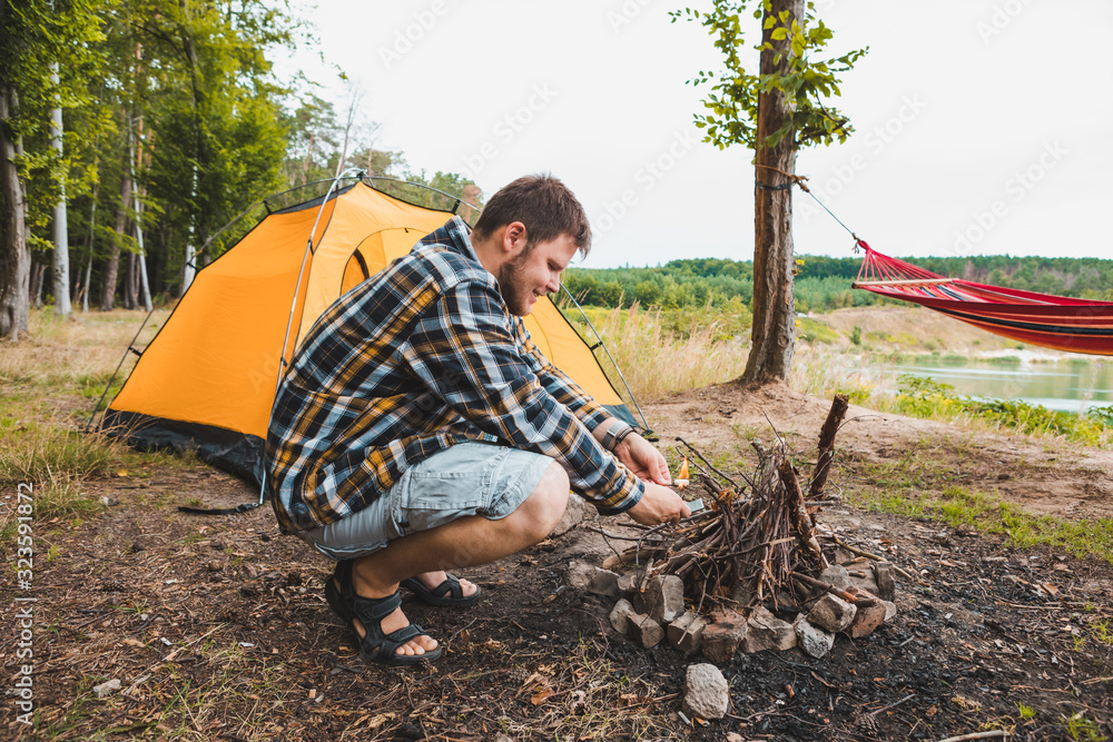 young strong man light a fire at camping site yellow tent on background young strong man light a fire at camping site yellow tent on background