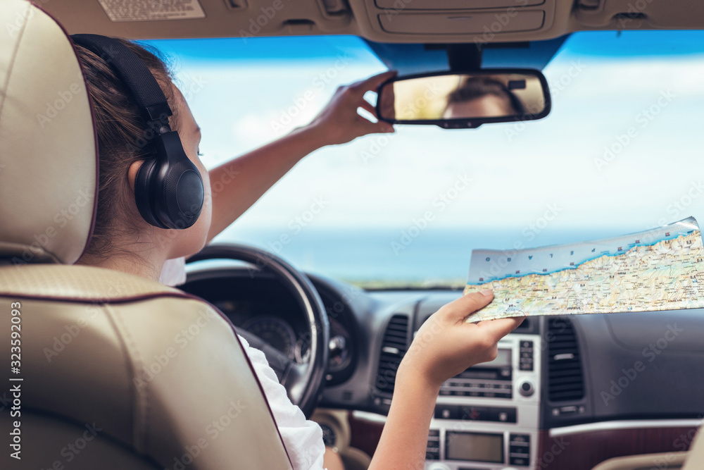 Woman driver in the headphones driving a car and looking at th map. Girl relaxing in auto trip traveling along ocean tropical beach in background. Traveler concept.