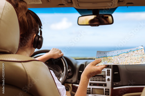 Woman driver in the headphones driving a car and looking at th map. Girl relaxing in auto trip traveling along ocean tropical beach in background. Traveler concept.
