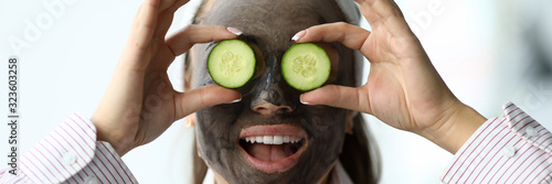 Woman makes gray clay facial mask at home. Put cut cucumber on her eyes like glasses. Spa home beaity skin mask make concept photo