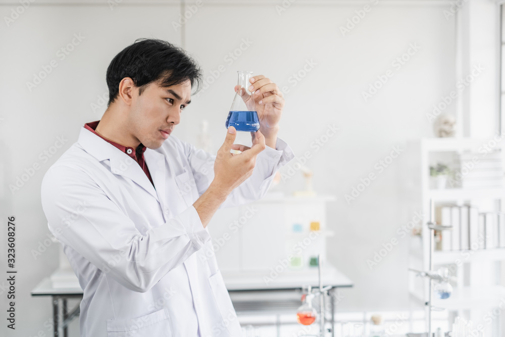 A male asian scientist staring seriously at a beaker with blue solution inside a laboratory setting. A male asian scientist staring seriously at a beaker with blue solution inside a laboratory setting.