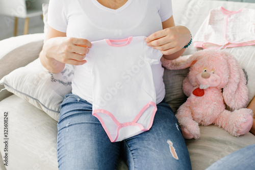 The expectant mother holds a baby white bodysuit with a pink canticle in her hands, and a pink stuffed toy hare is sitting next to her photo