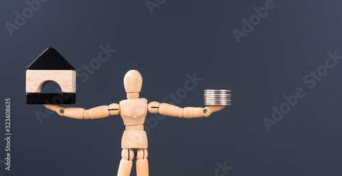 Wooden man comparing two things , hold handful of coins and black white mini house. Copy space for ad text. Concept of libra, percent, correct choice, property, credit, and money. Mannequin close-up
