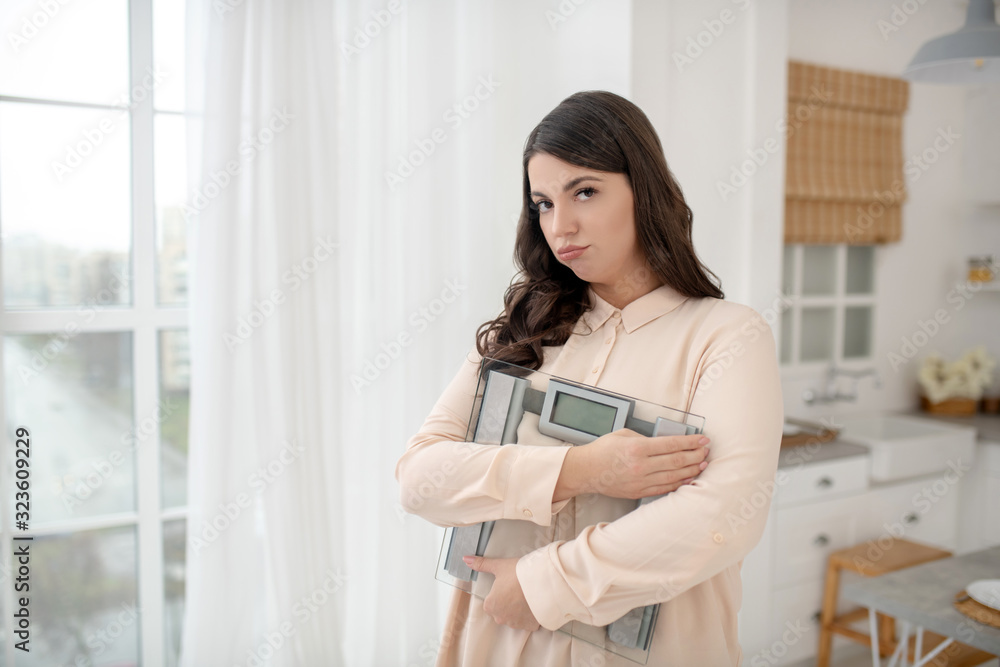Young woman in a beige blouse holding weigher and feeling stressed