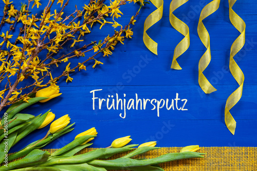 German Text Fruehjahrsputz Means Spring Cleaning. Yellow Spring Flowers Like Tulip And Branches. Festive Decoration With Ribbon. Blue Wooden Background photo