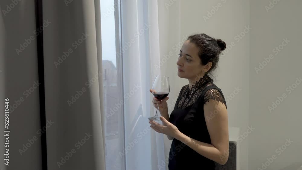 A lovely woman stands and drinks red wine from a glass glass by the window on a neutral gray background. Fashion portrait. Concept. Closeup. 4K. A lovely woman stands and drinks red wine from a glass glass by the window on a neutral gray background. Fashion portrait. Concept. Closeup. 4K.