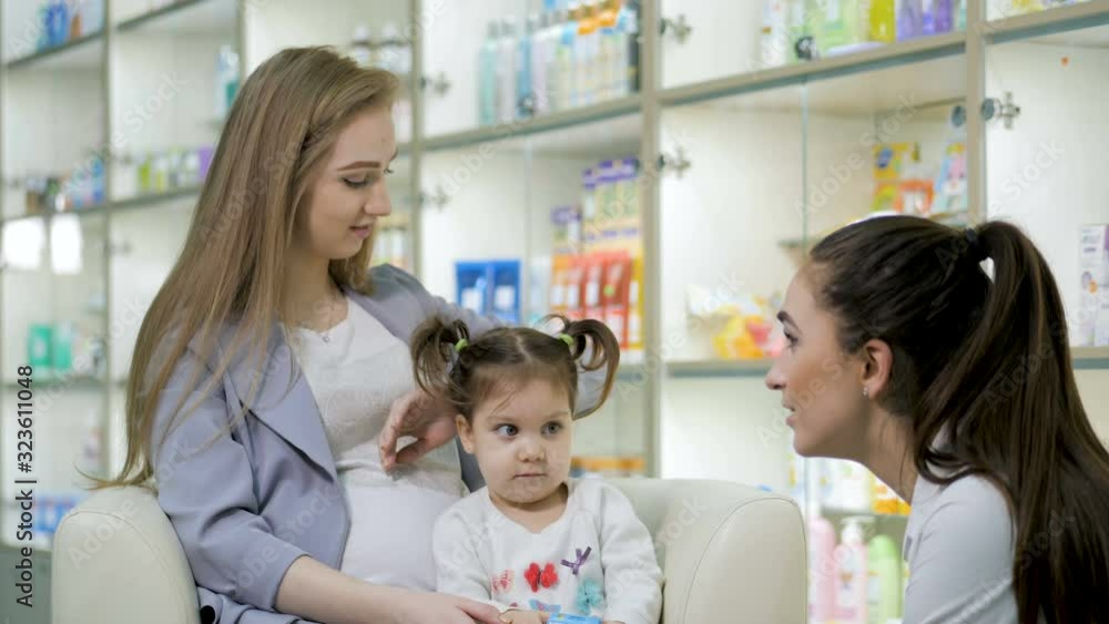 Young girl pharmacist giving advise to pregnant customer with cute baby girl. Pharmaceutical store concept Young girl pharmacist giving advise to pregnant customer with cute baby girl. Pharmaceutical store concept