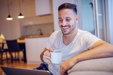 Handsome smiling unshaven caucasian man in pajamas sitting in living room with laptop in lap and drinking his fresh morning coffee.